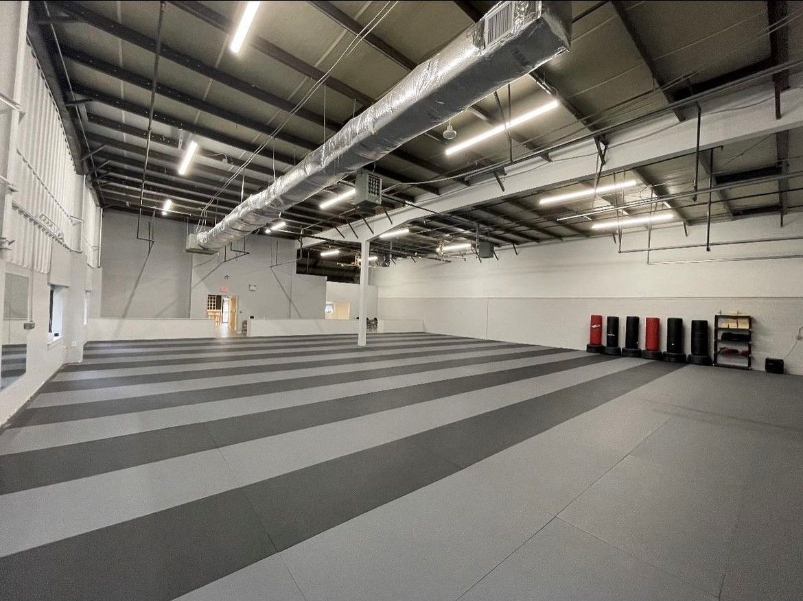 Large empty gym with gray and black floor mats, high ceilings, and training equipment.