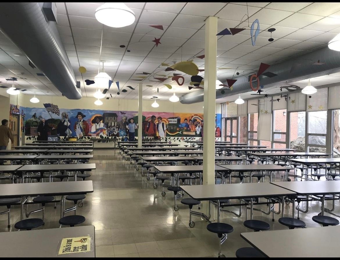 School cafeteria with long rows of tables, a mural, and ceiling decorations.