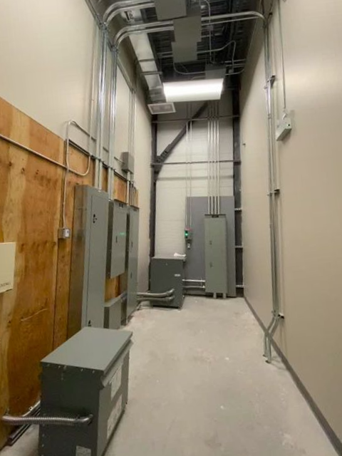 Narrow room with electrical equipment on the walls and floor, metallic conduits run overhead and along the walls.