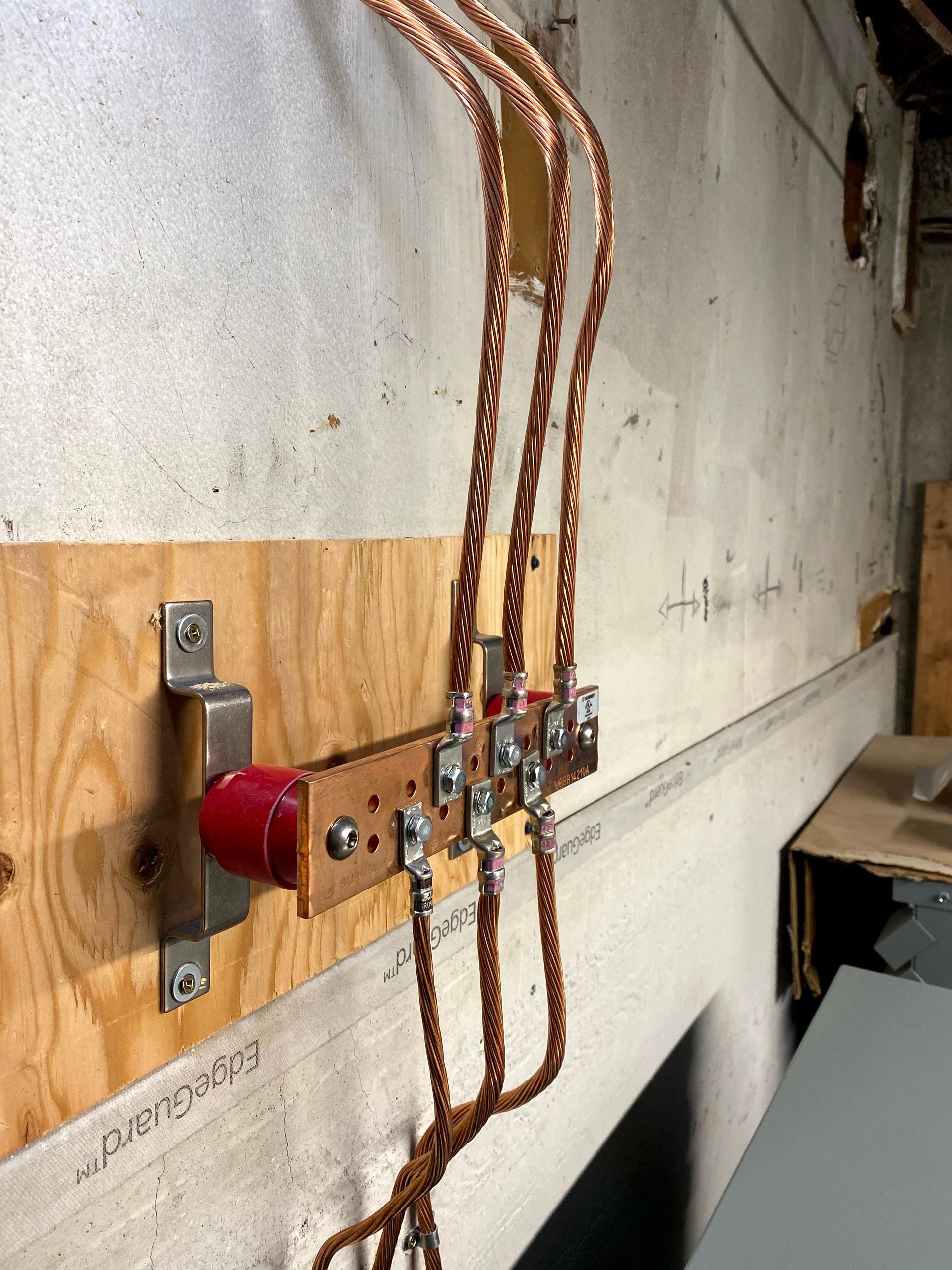 Copper electrical wires connected to a red switch on a wooden board attached to a concrete wall.