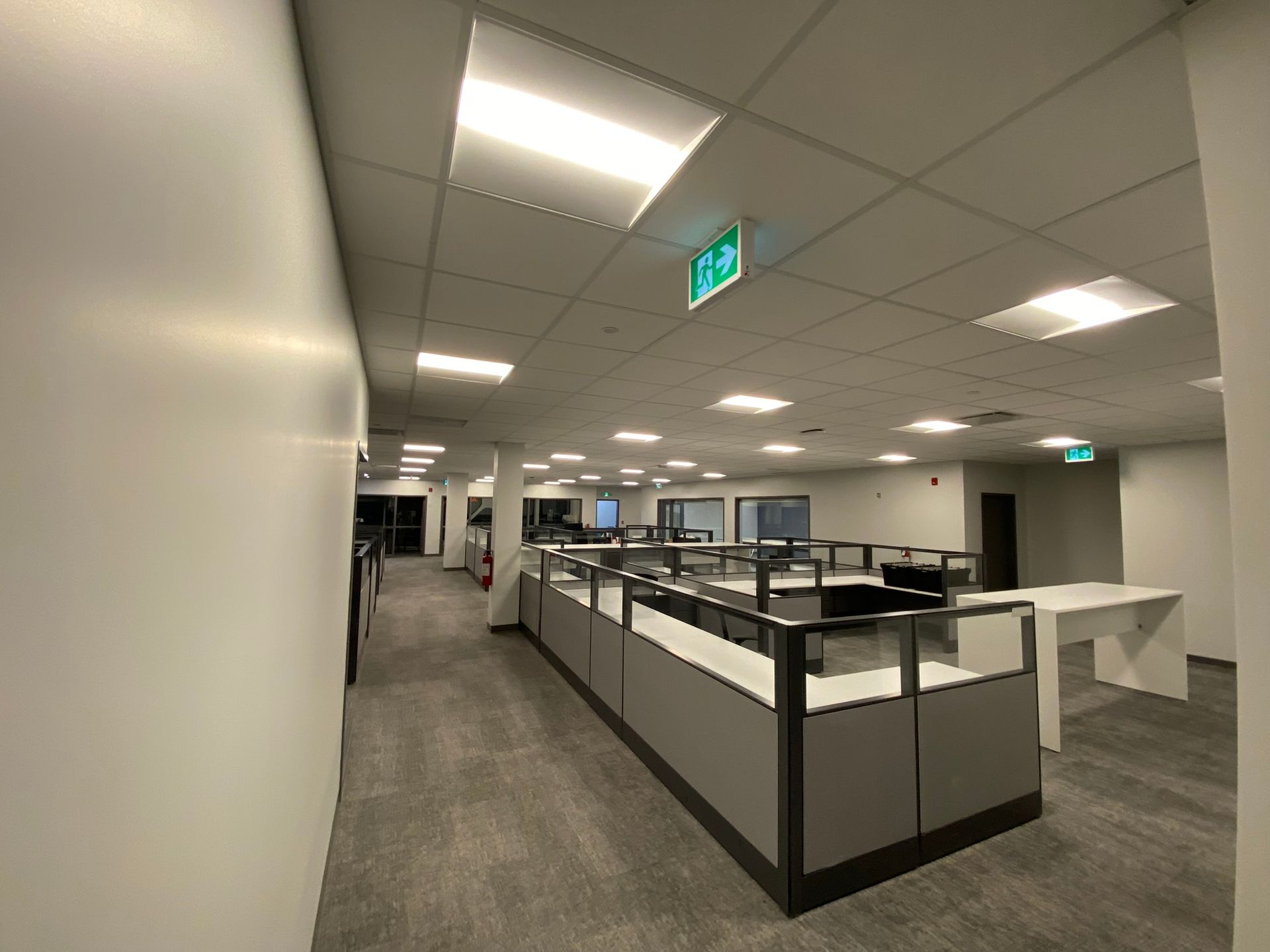 Office hallway with gray cubicles, recessed ceiling lights, and an exit sign.