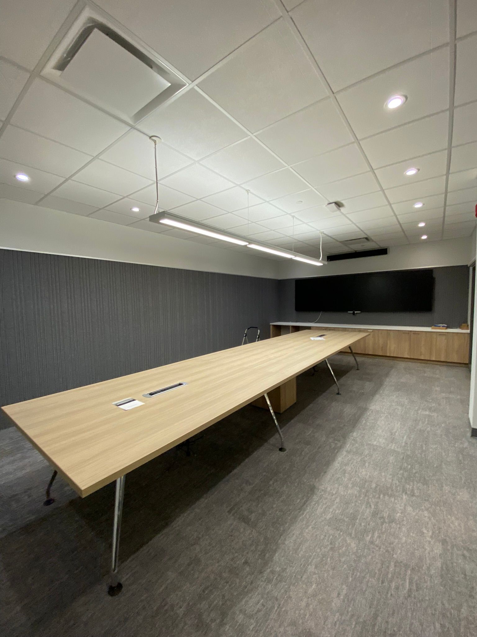 Long, light-colored conference table in modern room with large screen, neutral tones, and recessed lighting.