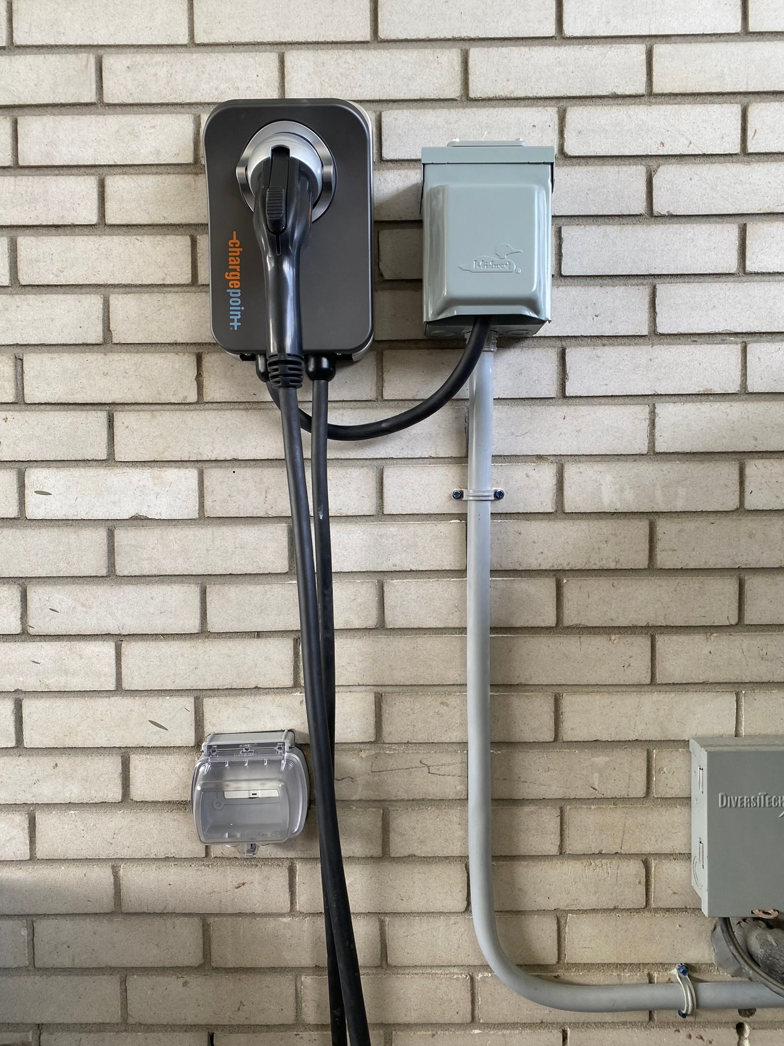EV charger and electrical box mounted on a brick wall. Gray and black components, gray conduit.