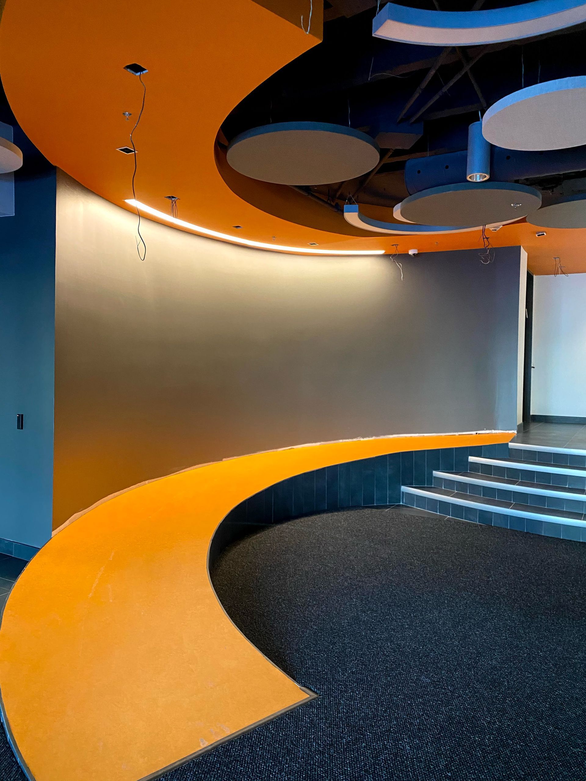 Orange curved pathway with a gray wall, blue ceiling, and carpeted floor.