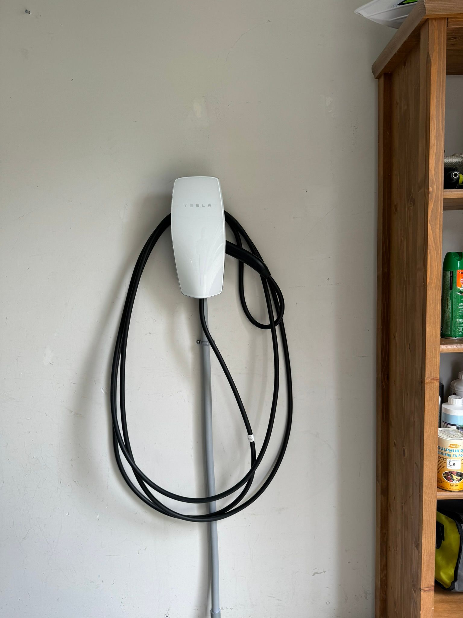 White Tesla charger mounted on a light gray wall with a black cable hanging down, next to a wooden shelf.