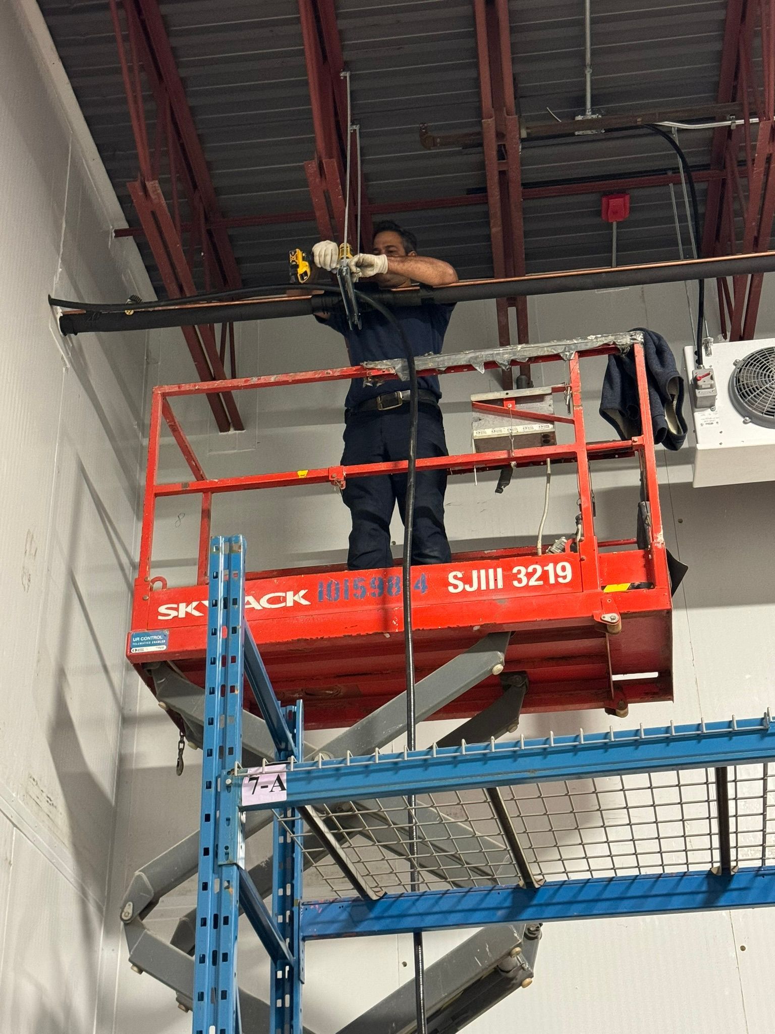 A person on a red scissor lift works on wires attached to a black track in a warehouse setting.