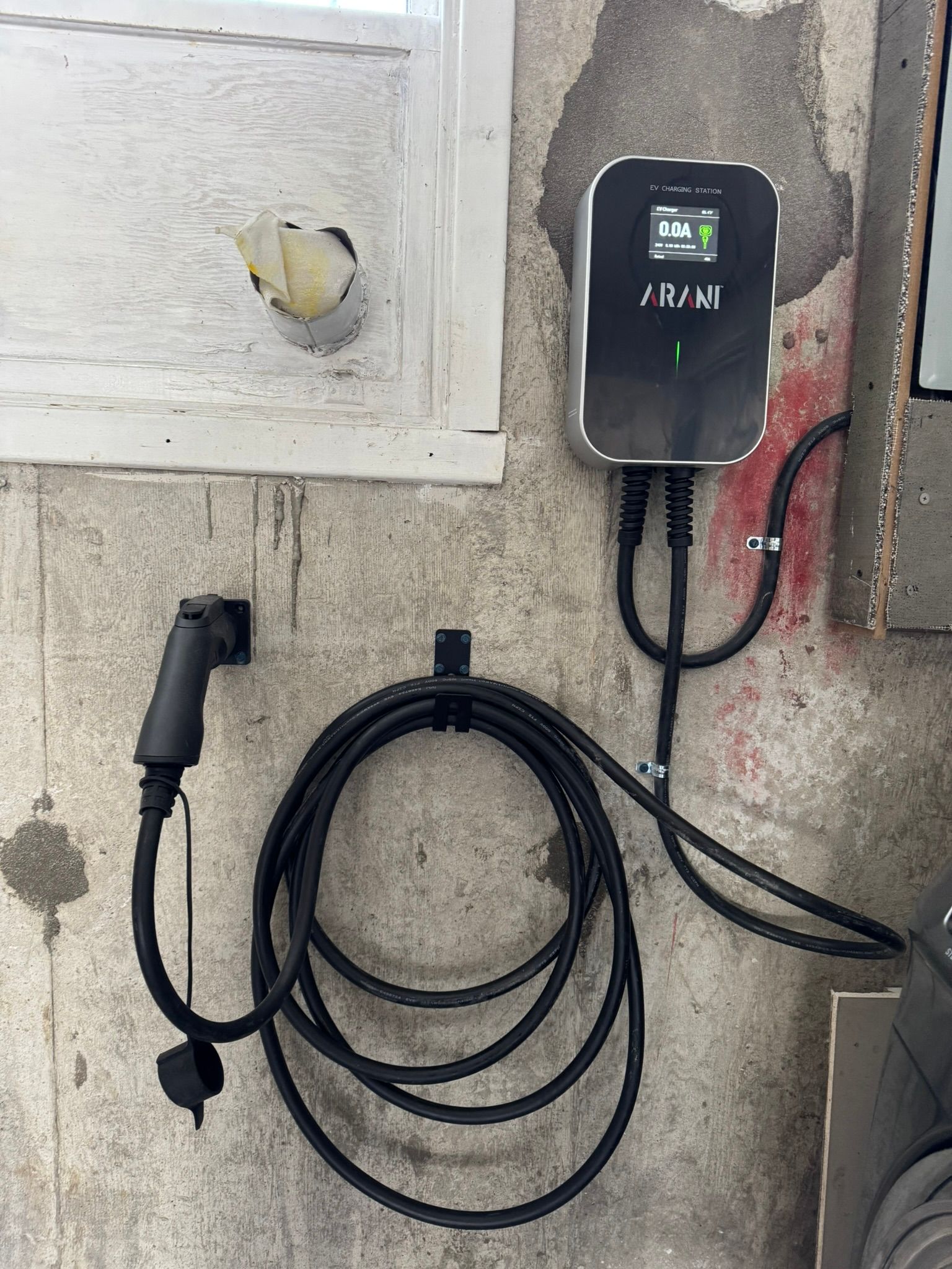 EV charger mounted on a gray, weathered wall; charging cable coiled, connector hanging.
