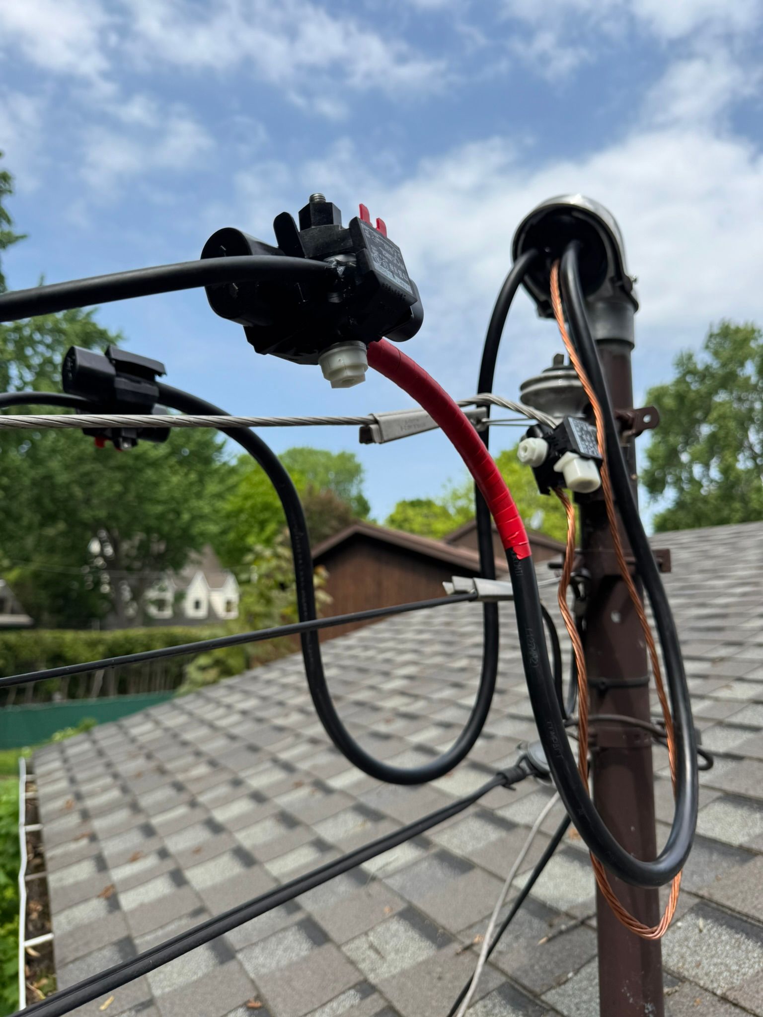 Electrical wires and components on a pole, mounted on a rooftop. Black and red cables, a fuse, and a metal enclosure are visible.