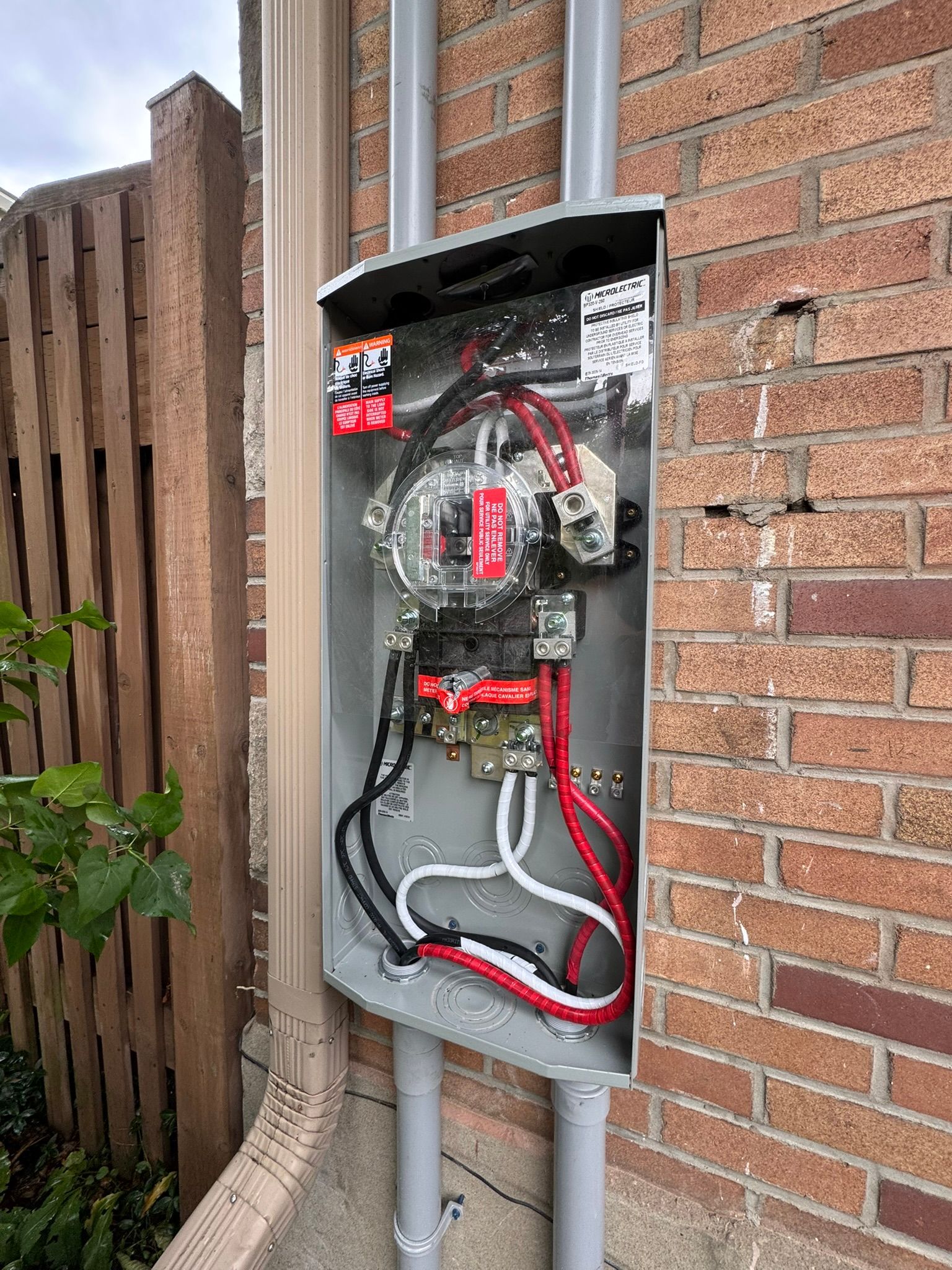 Electrical meter box mounted on a brick wall, wires visible inside. Gray box, red and black wires, and conduit.