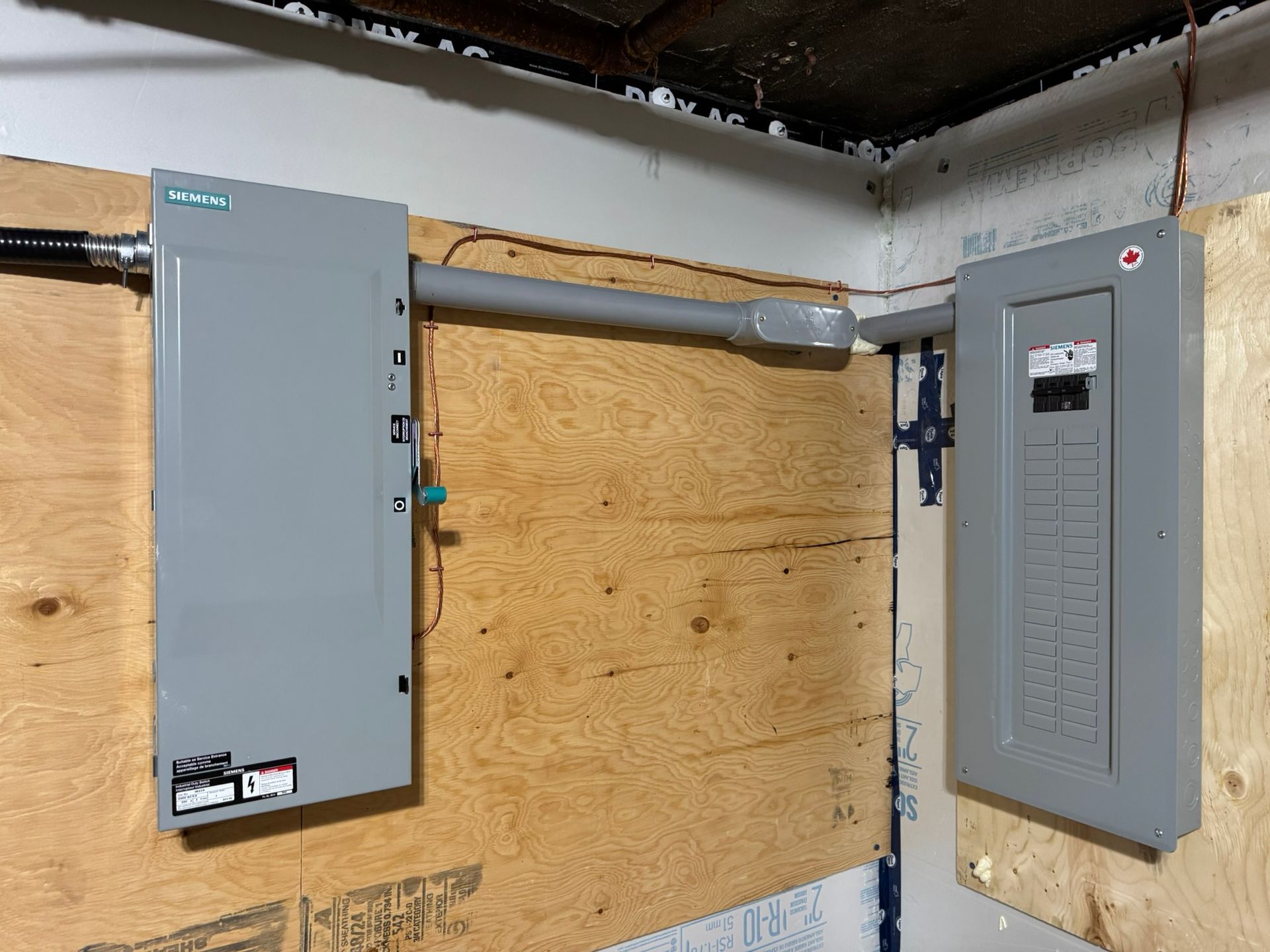 Electrical panel and subpanel connected by conduit on a plywood wall.