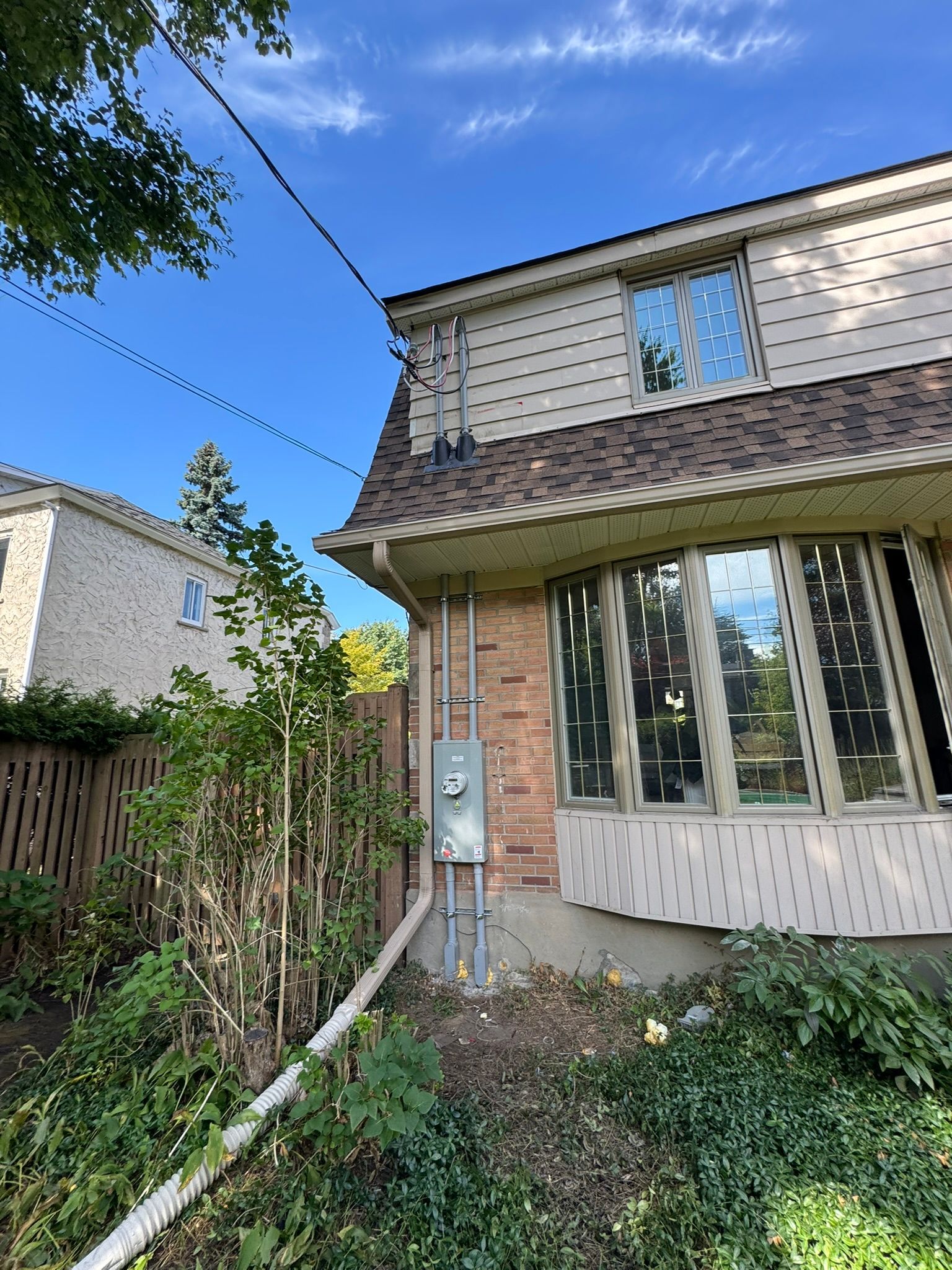 Exterior view of a two-story house with a brick and beige siding. Electric meter and wires are visible. Blue sky.