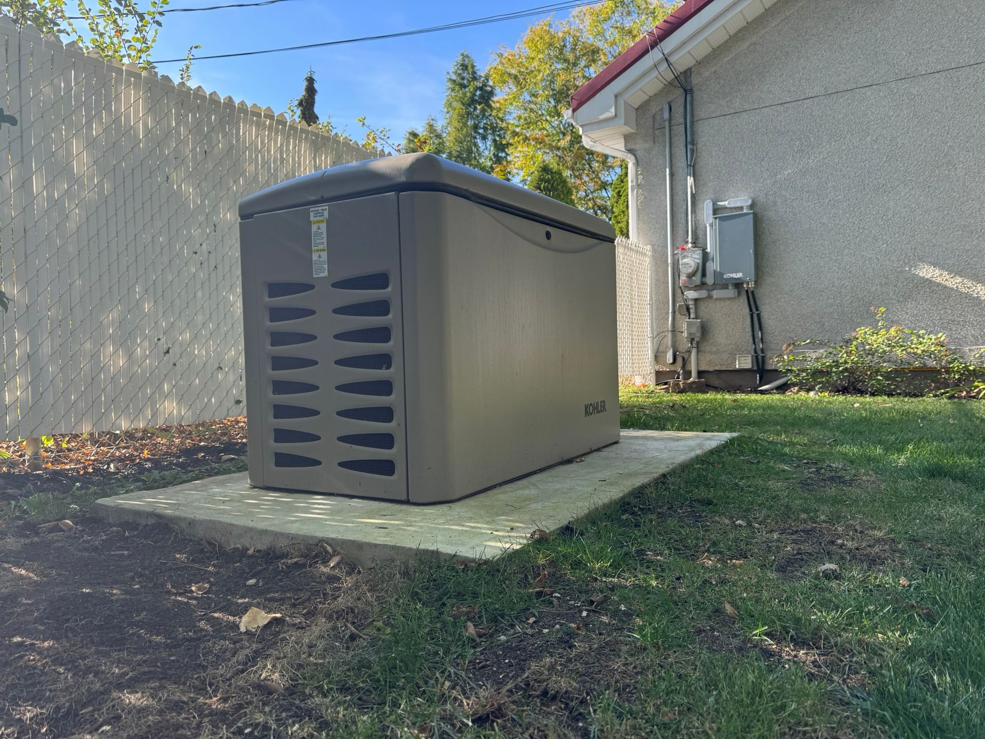 A standby generator on a concrete pad next to a house, with a white fence in the background.