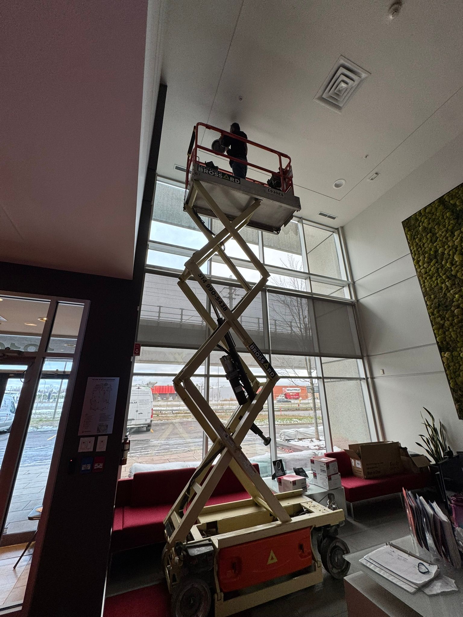 Person on a scissor lift working near a tall window. Building interior with red couch and green wall.