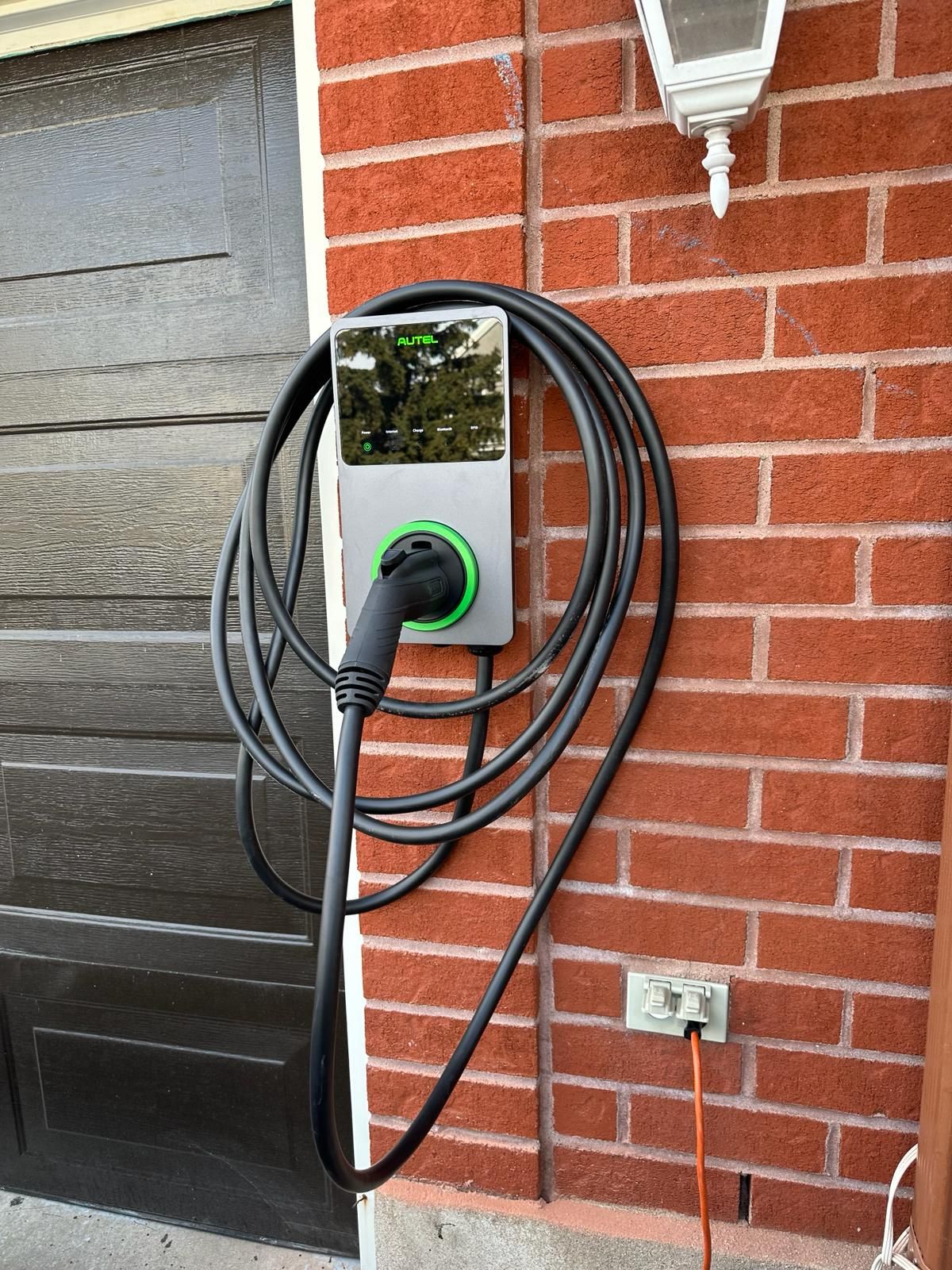 An electric vehicle charger mounted on a brick wall with a coiled black cable and plugged into an orange extension cord.