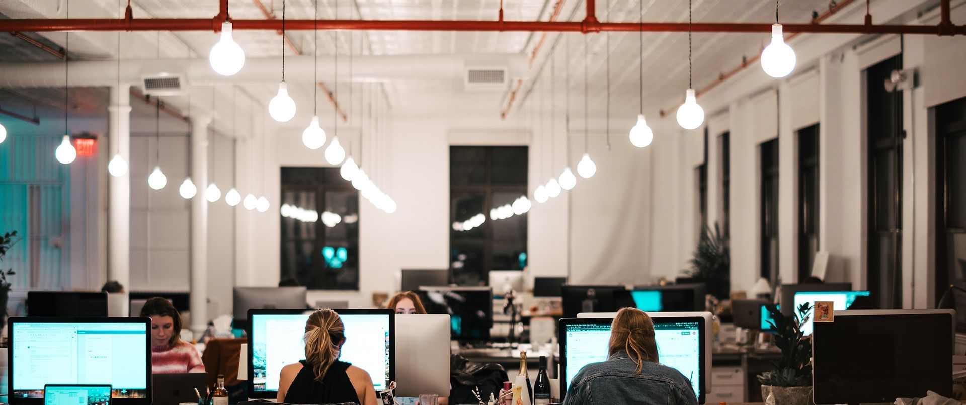 An open-plan office space with employees working on computers under hanging lights.