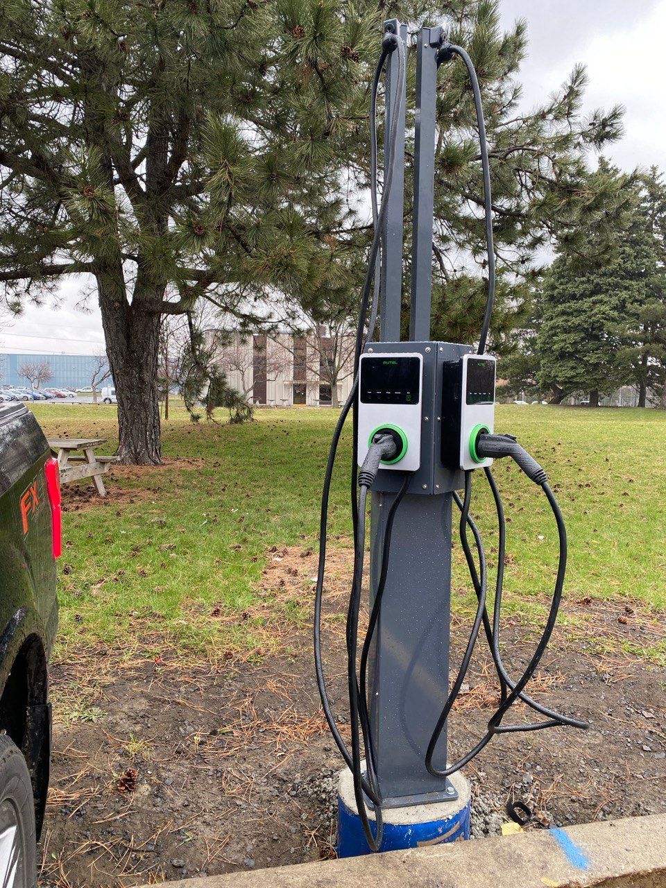 Borne de recharge pour véhicules électriques à deux ports, située dans un parc. Un véhicule est branché sur l'un des ports.