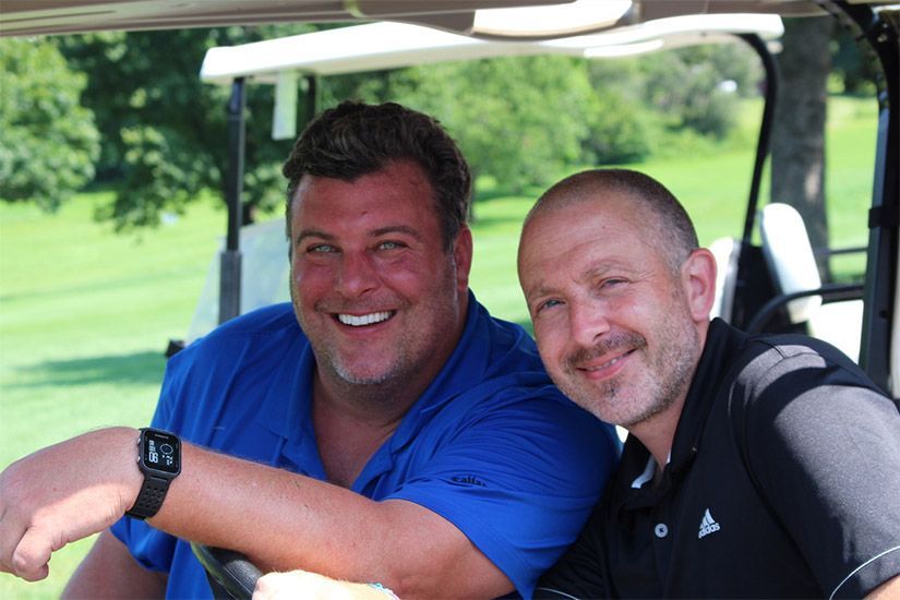 Two men are sitting in a golf cart and smiling for the camera.