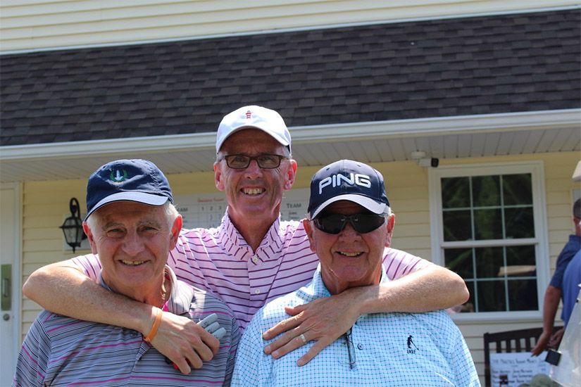 Three men are posing for a picture in front of a house . one of the men is wearing a ping hat.