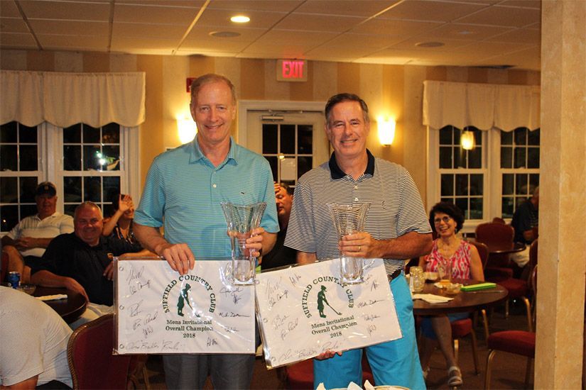 Two men holding trophies in a room with an exit sign in the background