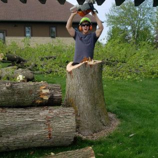 A man is standing on a tree stump holding a chainsaw over his head.