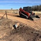 An orange skid-steer loader parked on a dirt road near an exposed black plastic culvert pipe in a rural field.