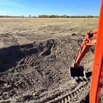 An orange excavator arm and bucket sit at the edge of a deep, dirt-filled pit in a dry, open field under a blue sky.