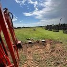 An orange excavator arm overlooks a dug-out patch of dirt and grass in a large, sunny, open field.