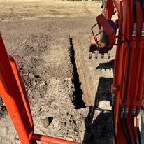 A view from the cab of an orange excavator, looking down at a freshly dug trench in a dirt field.