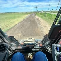 First-person view from inside a skid steer looking out over a leveled dirt driveway leading to an open gate.