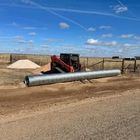 A skid steer loader lifts a long, corrugated metal culvert pipe on a dirt road beside a pile of gravel under a blue sky.