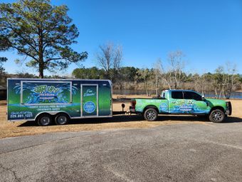 Green truck towing a matching trailer with business logos, parked near a body of water.