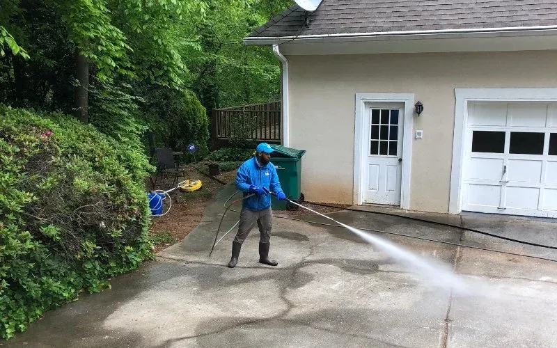 Person in blue washing driveway with pressure washer in front of a garage.