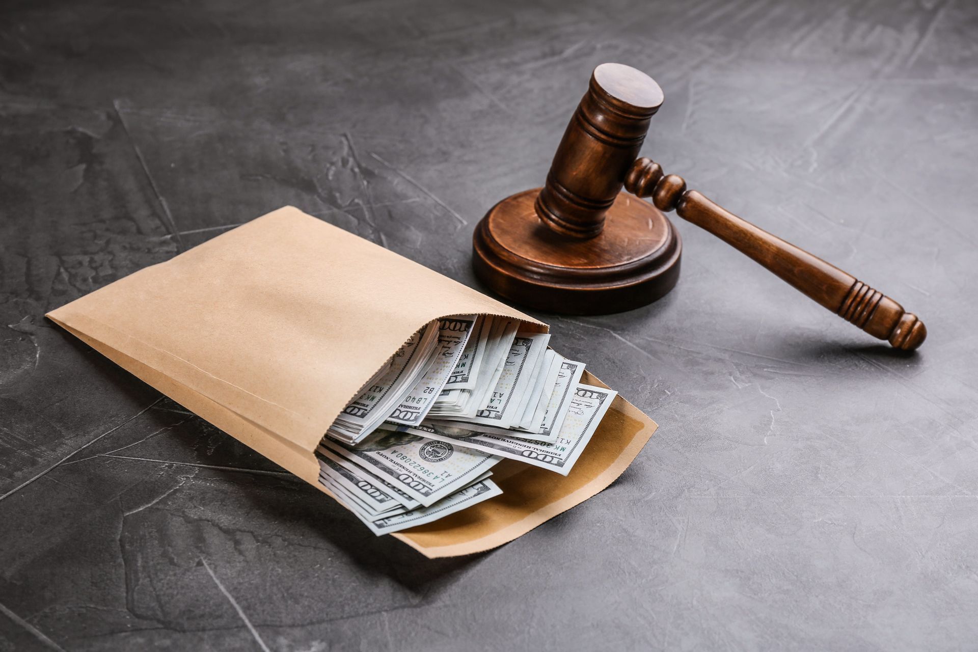 A stack of US dollar bills in a tan envelope sitting on a gray surface next to a wooden judge’s gavel.