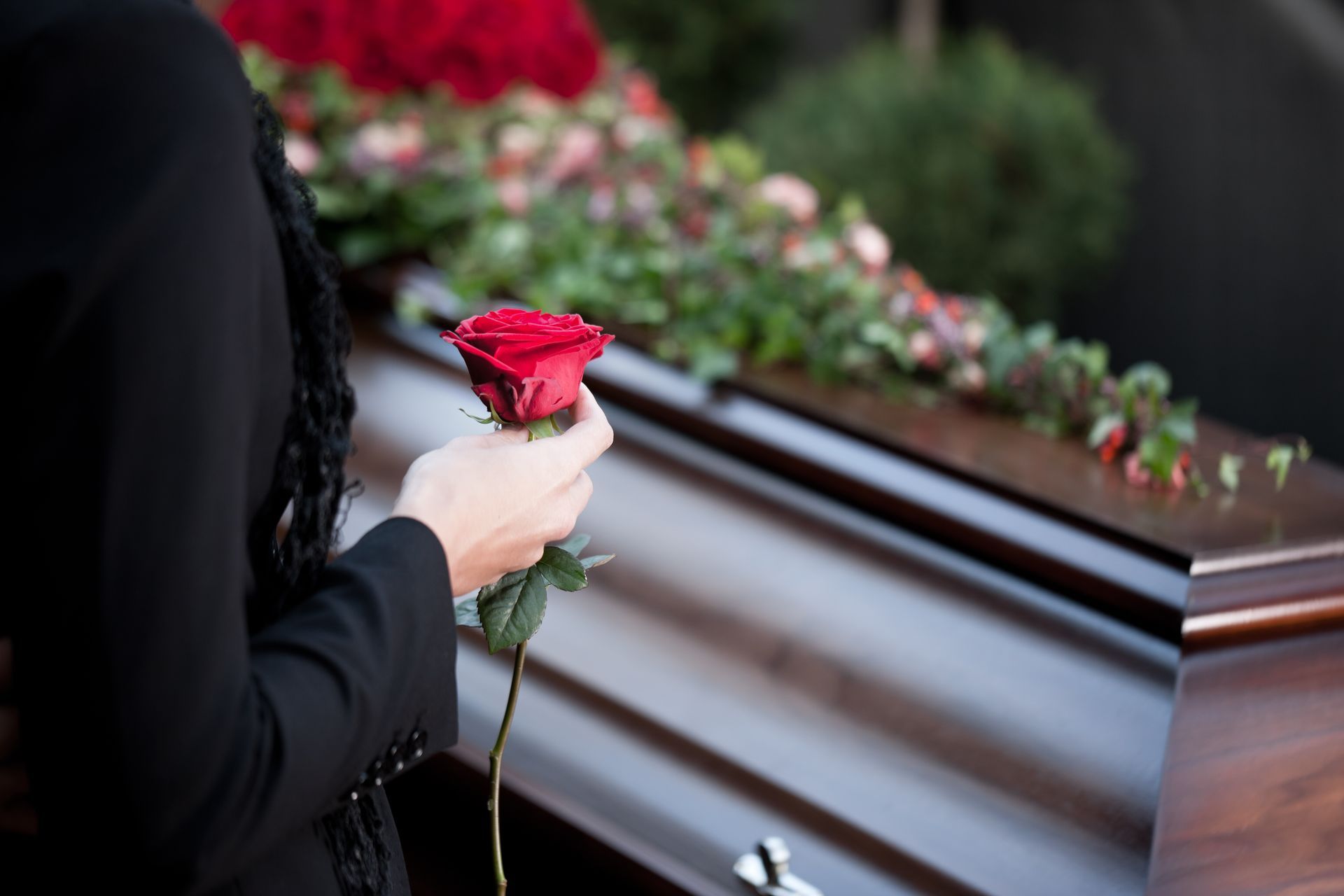 A woman is holding a red rose in front of a coffin.