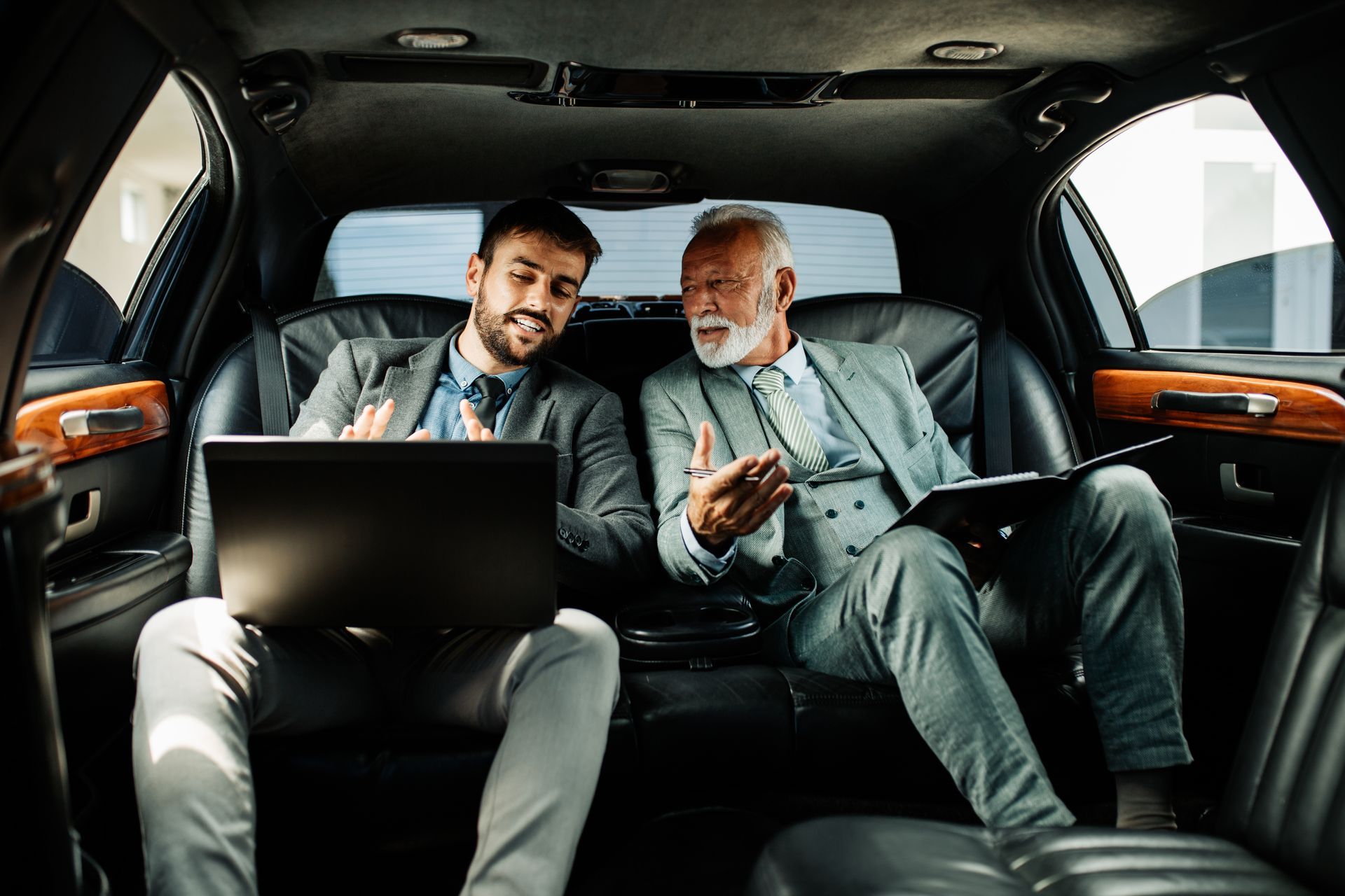 Two men are sitting in the back seat of a limousine looking at a laptop.