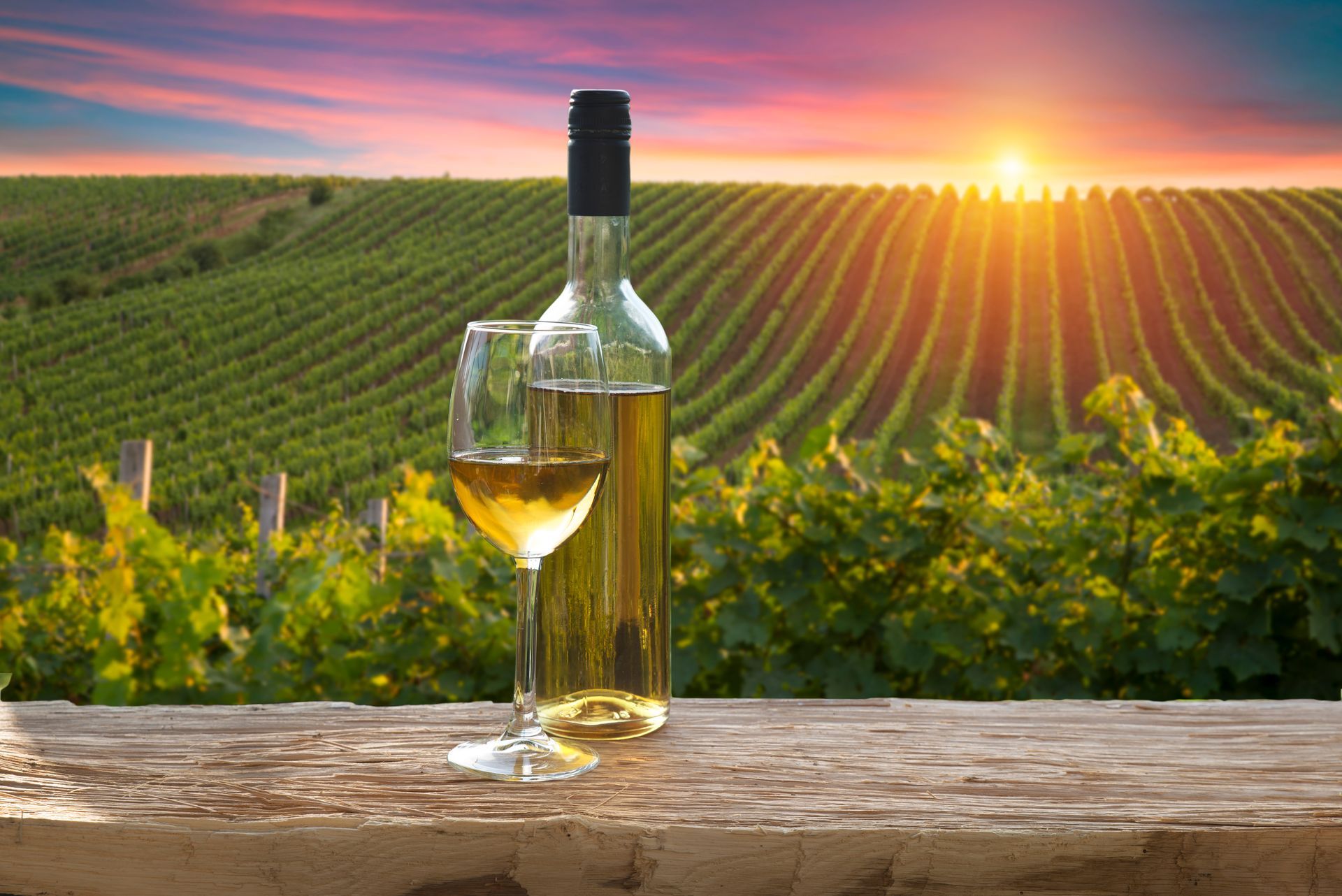 A bottle of wine and a glass of wine are on a wooden table in front of a vineyard.