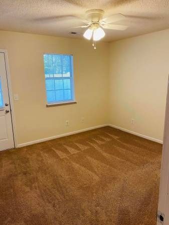 Empty room with tan carpet, beige walls, small window, ceiling fan, and door.