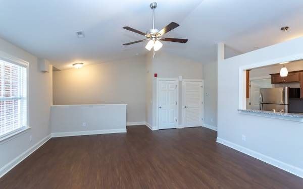Empty living room with wood flooring, neutral walls, and a view into a kitchen.