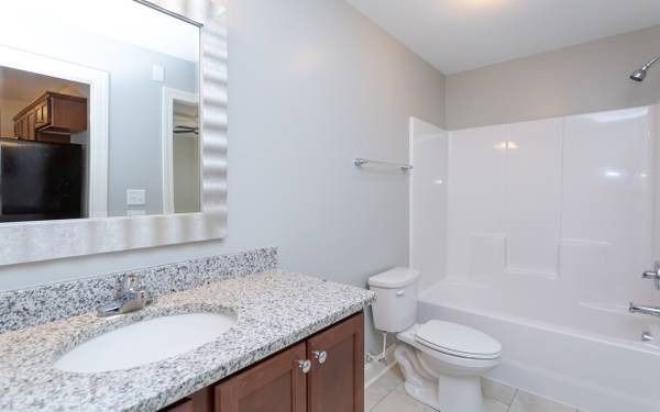 Bathroom with a white tub, toilet, and vanity with granite countertop. Mirror reflects a kitchenette.