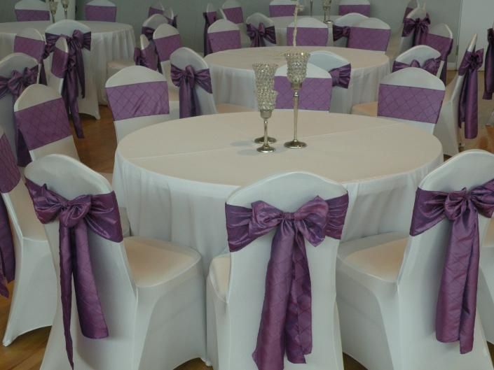 Round tables with white chairs tied in purple bows at a decorated banquet hall.