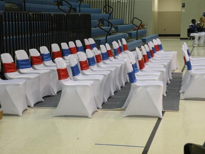 Rows of white-covered chairs with red, white, and blue sashes in a gymnasium