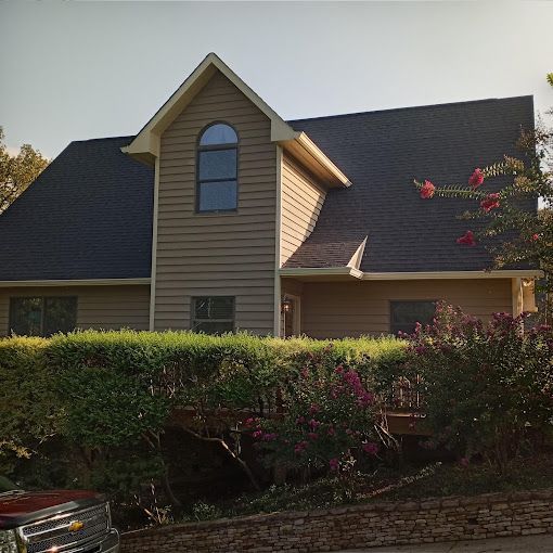 A large house with a black roof is surrounded by bushes and flowers.
