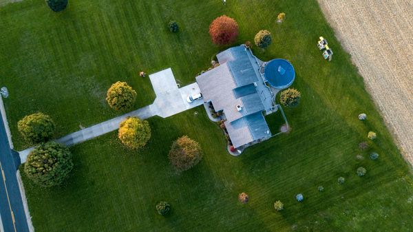 Aerial view of a house with a driveway, pool, and green lawn, adjacent to a field.