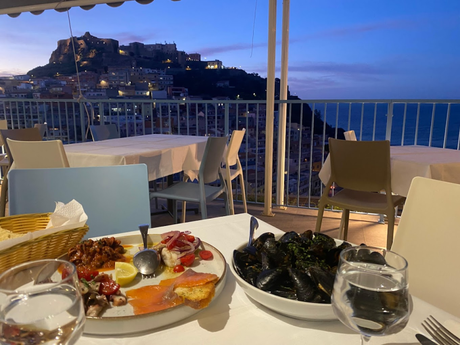 Una cena a base di frutti di mare e cozze servita sulla terrazza di un ristorante al crepuscolo, con vista su una cittadina costiera e sul mare.