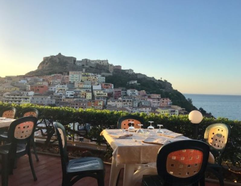 La terrazza di un ristorante con tavoli e sedie si affaccia su una cittadina costiera costruita su una collina in riva al mare, al tramonto.