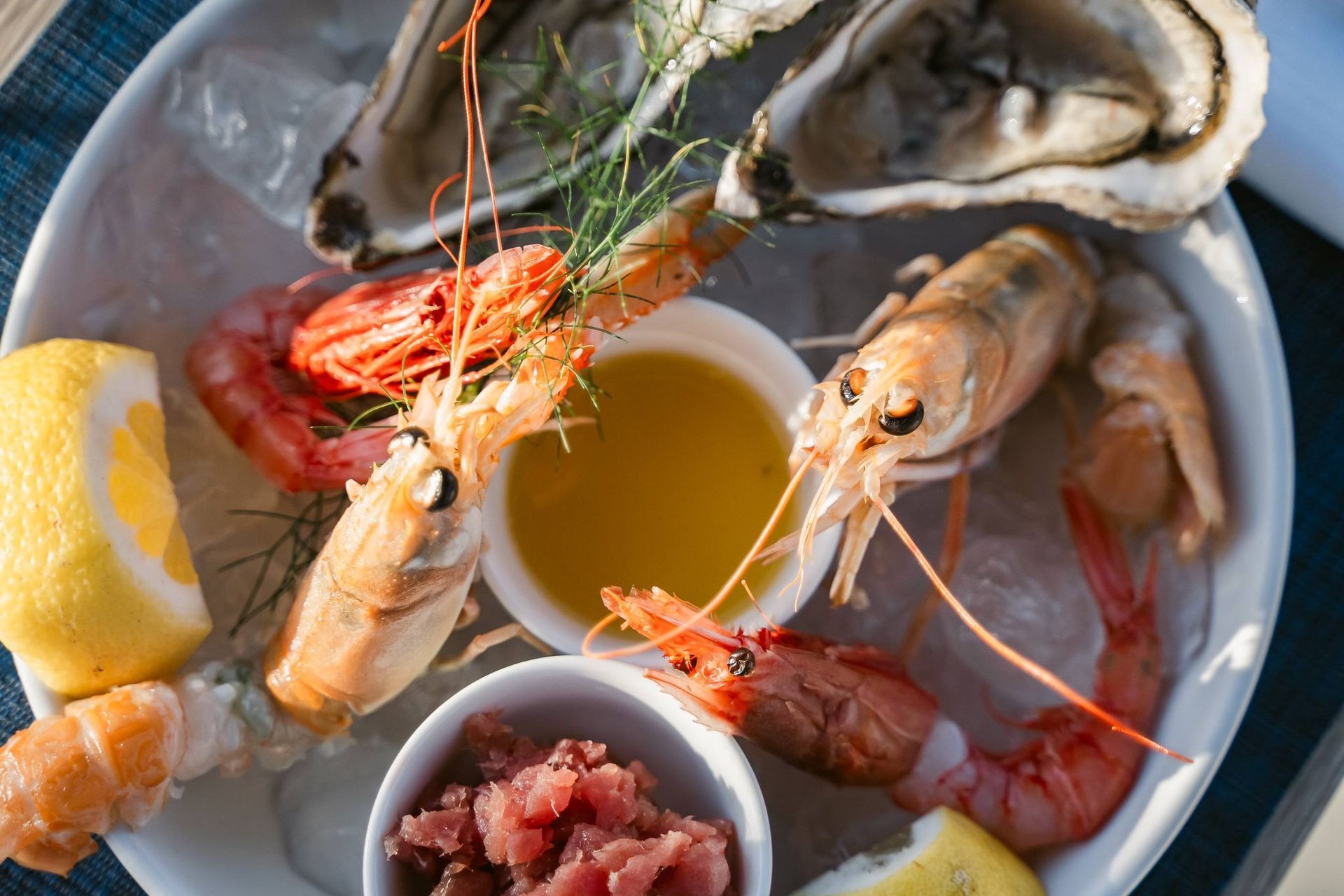 Un piatto di frutti di mare con ostriche, gamberi crudi e una porzione di tonno tritato, disposto su ghiaccio con spicchi di limone e salsa.