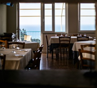 Interno del ristorante con tavoli apparecchiati con tovaglie bianche, sedie in legno e un'ampia vetrata con vista sul mare.