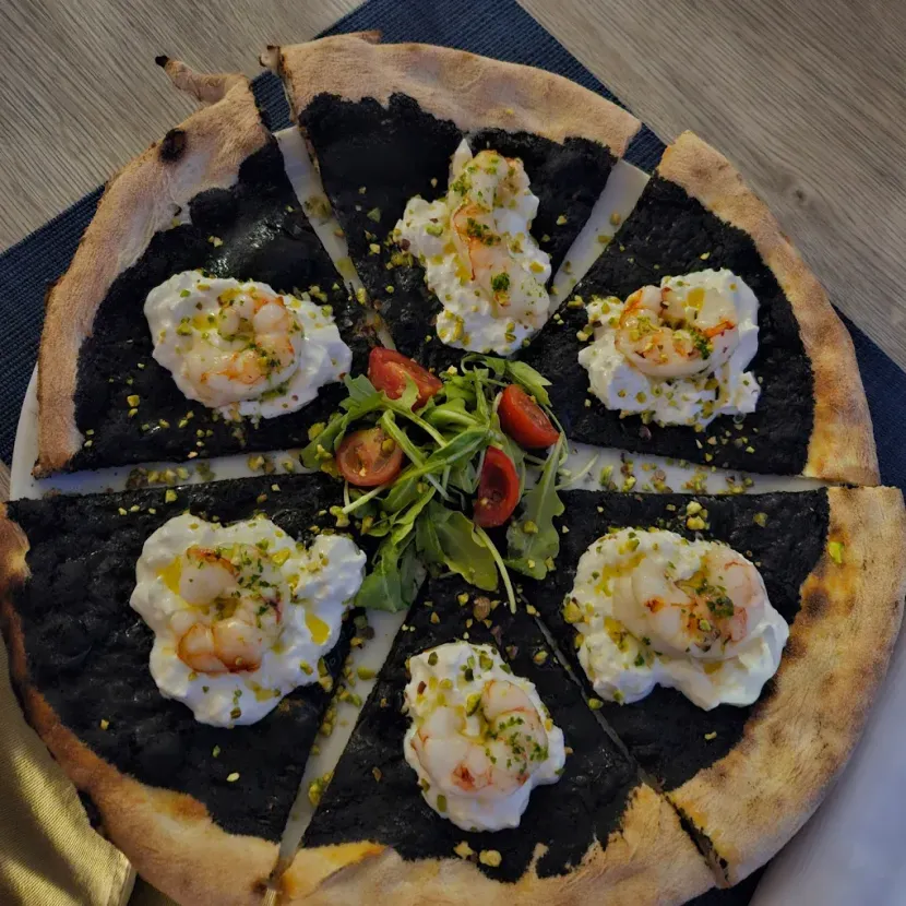 Una pizza con crosta nera, condita con formaggio bianco, gamberetti, pistacchi e guarnita al centro con rucola e pomodorini.