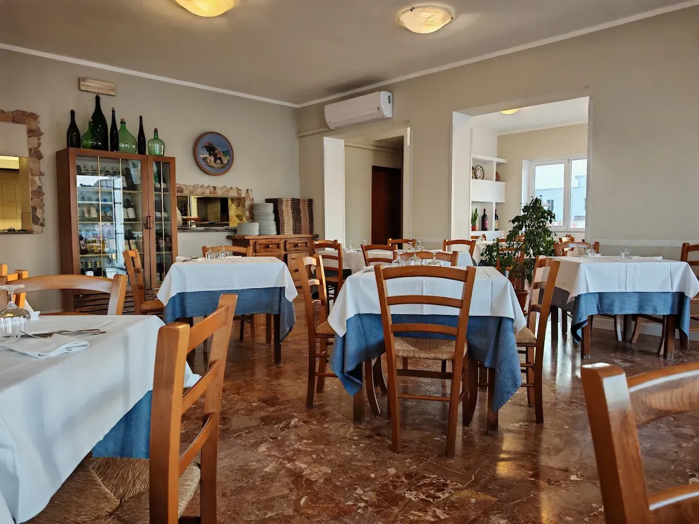L'interno vuoto di un ristorante con sedie di legno, tavoli coperti da tovaglie bianche e blu e una vetrina.