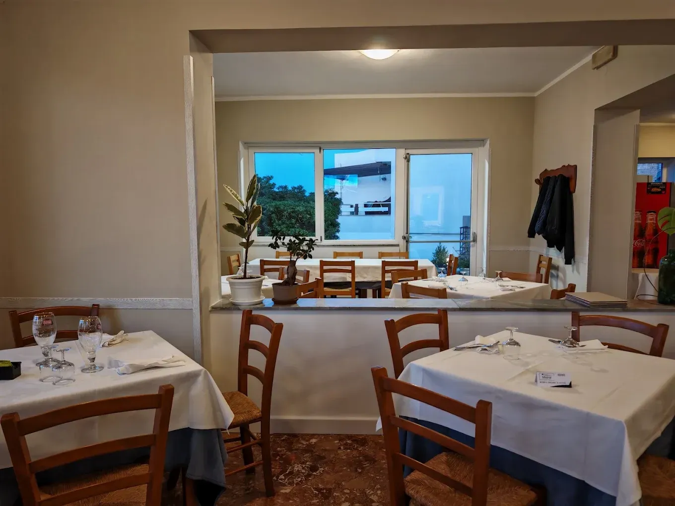 In un ristorante, due tavoli da pranzo con tovaglie bianche e sedie di legno, affacciati su una finestra e circondati da piante d'appartamento.